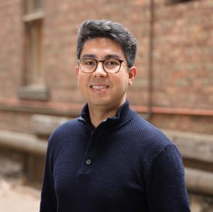 Elbaite CEO and co-founder Mortaza Tollo stands in front of a brick wall, he is wearing a dark blue knitted jumper and glasses