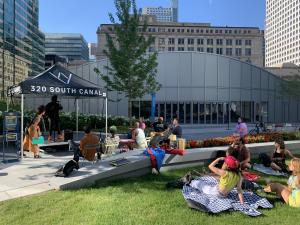 Approximately a dozen people in summer clothing are seating outdoors in folding chairs or on blankets on a grassy lawn.  They are looking in the direction of a man who is standing on a small stage with a navy canopy overheard that reads "320 South Canal."