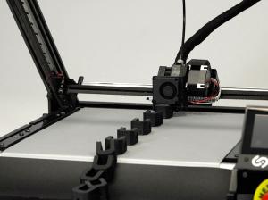 The One Pro 3D belt printer, which makes small components out of PETG material, all lined up and starting to gather at the end of the conveyor belt