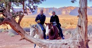 Chip Walter and Cyndy Mosites sit astride white and brown horses behind a tree with trunk and branches shaped like a picture frame.