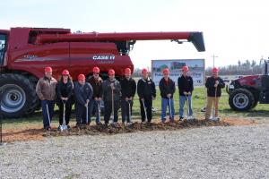 Ten Redline Equipment employees with hard hats hold shovels as they break ground for a new facility in Logansport, Indiana. A Case IH piece of farm equipment provides the background.