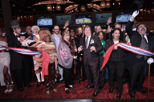See image: A photo of various people, employees of The Temporary and entertainment dressed as American presidents, circle around a red, white, and blue ribbon to represent the American Place's logo. The ribbon has been cut and the guests in the photo are