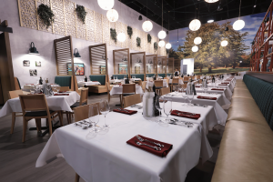 The photo is at an angle displaying multiple dining tables, as well as booth arrangements, with silverware,  wine glasses, red napkines, and white linens. The background shows a wall painted with a tree in front of a forest, and the left facing wall has v