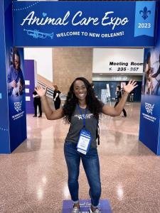 Black woman standing in a conventional hall under a sign for the Humane Society Animal Care Expo. She is smiling and pointing to the sign above her head with both hands.