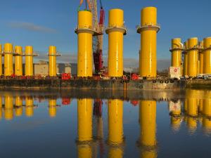 An array of bright yellow transition pieces are seen. Behind one is a crane which is connected to the transition piece. An orange hue colours the horizon, which contrast with the clear blue sky.