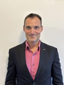 This photo of a smiling man wearing a dark colored suit jacket and pink dress shirt is Mr. Sandro Di Simplicio, the surgeon at the Royal Victoria Infirmary who performed the implant. He is looking directly at the camera, is by himself in the photo with a 