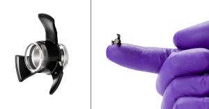 This photo is a composite of two images. On the left is an enlarged view of the SING IMT device, which is comprised of a circular center glass piece surrounded by three black silicon "arms". The image on the right is a real-size photo of a hand covered in