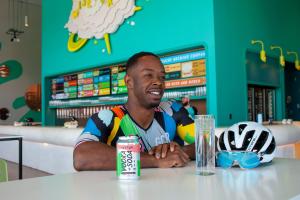 A photo of Derek Hosey wearing his brightly colored The Set Up cycling kit with cans of The Set Up in front of a teal wall.