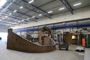 A man is tanding within the test facility encompassed by a large rounded steel structure, which will form part of the new man is send-of-line test rig designed for offshore powertrains