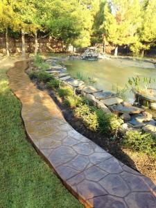 Photo of a cobblestone stamped concrete walkway next to a pond with a fountain in the middle and a garden bed of purple flowers.