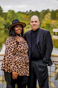A man in all black suit and woman in a leopard print shirt with black pants standing on a lake dock with fall color trees in the background