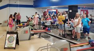 Wide shot of several people smiling at the bowling alley. There are people with and without disabilities and one in a wheelchair