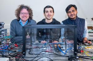 Three researchers in casual clothes stand behind an optical table in the laser laboratory of Leibniz Universität Hannover, in front of them is a photonic chip in a glass box. Copyright: Sonja Smalian/PhoenixD