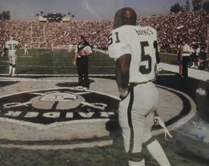 Rodrigo Barnes holding helmet and gazing at the field on the sidelines with John Madden in background as well as other coaches during Oakland Raiders game.