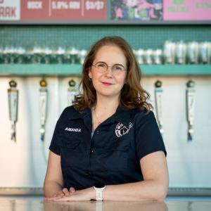 A photo of a woman (white with brown hair, navy shirt) standing behind a bar in front of several beer taps at WISEACRE's Memphis headquarters.