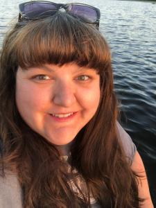 Image show Tasha Ferris who has long brown hair, hazel eyes, and a blue shirt. Lake Jean at Ricketts Glen State Park can be seen behind her.