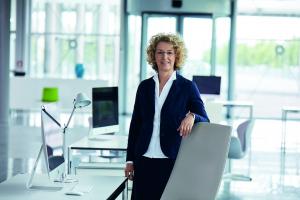 Product Manager Michaela Imbusch (woman with curly blond hair and a dark blue woman's suit) standing in a bright office besides her desk chair.