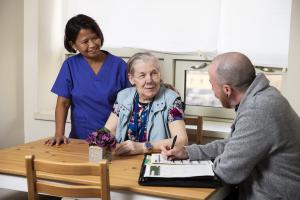 A Home Care patient is seated at her kitchen table in her home, while a research coordinator who is also seated takes notes. Behind the patient, a Home Care nurse is standing and smiling, as she gently pats the patient's shoulder.