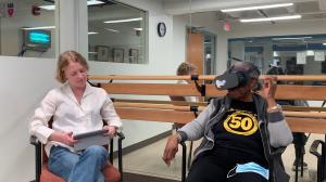 Two women sit smiling in chairs in front of a wall-sized mirror. They are talking. The woman on the right is wearing a VR headset and appears to be moving her hands to emphasize what she is saying.