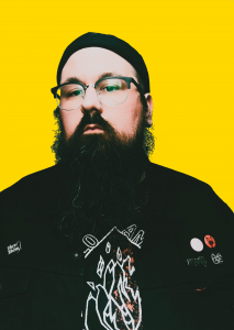 Author in black jacket, shirt and hat; black beard and glasses, stands against yellow background