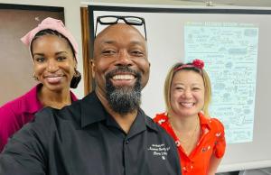 Dr. Cassandra Bolar, Dr. Rodney Washington and Rio Holaday stand together and smile in front of visual aid for Community Listening Session in Monroeville, AL