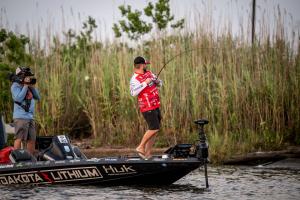 Brock Mosley, professional angler, standing in his boat powered by Dakota Lithium during the Bassmaster Elite tournament. He holds a fishing rod with a fish on the line, showcasing his skill and the performance of Dakota Lithium batteries. The boat promin