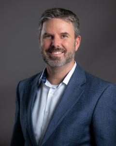 Ambassador Enterprises Chief Legal Officer Jeremy Reidy portrait wearing white dress shirt and glen-plaid sport jacket on gray background