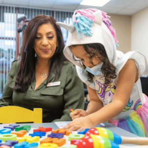 Young girl in mask working with blocks and Variety team member is watching and helping