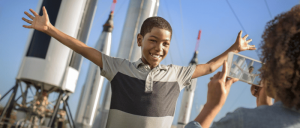 Boy standing in front of rocket as mom takes a photo of him smiling