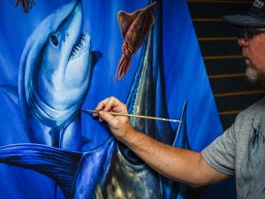 Photo depicts artist, Dennis Friel, painting an ocean scene containing a shark, swordfish, and squid.