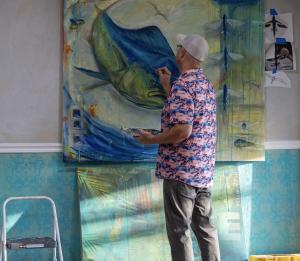 Image depicts artist, Derek Redwine, standing in front of and finishing his latest painting.