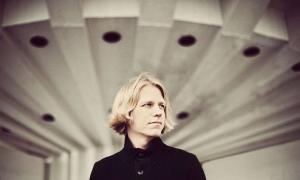 mysterious attractive Blond man with shoulder length hair in dark coat stands in front of futuristic building
