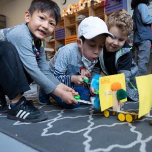 Three children kneel on the ground while playing with robots they've built.