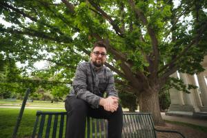 Chris Angel sitting on the top of the back of a park bench, hands clasped in front of them. They are slightly smiling at the camera with a grassy field behind them. A large tree towers over from behind, offering shade.