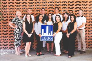 The 1836PM team grouped together in front of an orange wall holding a sign with the 1836PM logo.