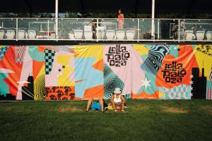 Two festival-goers lounging on the grass in front of a colorful Lollapalooza mural.