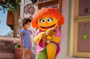young girl next to Julia the autistic muppet, who is knealing