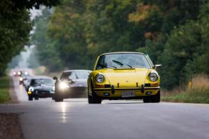 Vintage cars being led by a Porsche down a rural road surrounded by trees