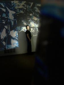A pale white woman with dark hair stands in profile in projected video of nature in a lobby space at Audium, a venue for sound art installations