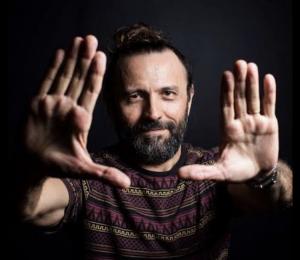 Photography of Halder Gomes. White man with black hair and long bear. His hand are open in front of his face. 