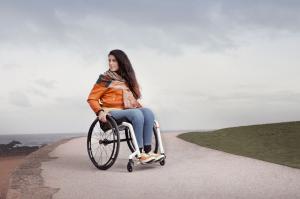 A woman with long brown hair, an orange coat and jeans sits in a manual wheelchair on a road next to the sea