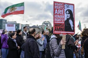 A group of demonstrators march in the street in solidarity with protesters in Iran. Towards the background, someone is holding an Iranian flag. In the foreground on the right side, someone holds a sign with a photo of Mahsa Amini, and the words next to it