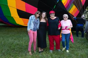 Four women stand in front of a rainbow-colored hot air balloon, with an operator in the background preparing it for takeoff.
