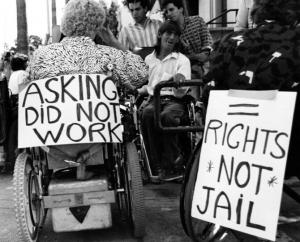Vintage photograph of wheelchair users with protest signs that read, "asking did not work," "= rights no jail."