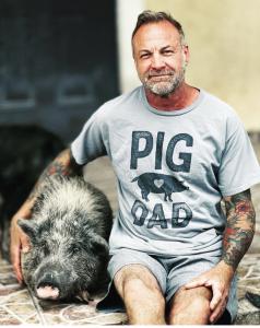 Little Bear Sanctuary Founder and Director Christopher Vane wearing a tshirt that reads Pig Dad, sitting on his porch next to one of his wards, a pig named Chubs