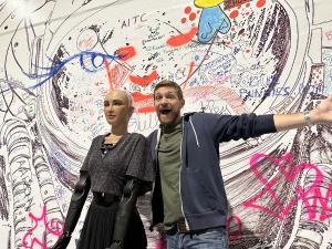 This image depicts Sophia the Robot and Aaron Vick standing together in front of the Tezos Commons BREATHE! communal Guinness World Record attempt, which is a large canvas with artwork alongside the convention wall