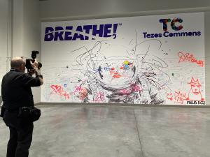 This image depicts a photograph taken of the entire Tezos Commons BREATHE! Communal Guinness World Record attempt, which is a large canvas of artwork displayed along the wall of the convention. A man is seen on the left-hand side of this image taking a ph