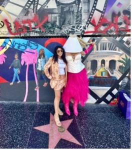 Farrah Mechael & Shitty Princess posing on Hollywood Boulevard Walk of Fame. Shitty Princess wears a pink tutu, white shirt, and white lampshade on her head. Farrah Mechael wears a light set of a shirt & skirt along with a white shirt. The mural behind them is vibrant.