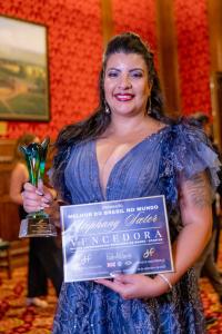 Mulher com cabelos longos escuros, sorrindo, maquiada e usando batom vermelho, segura na mão direita um troféu feito murano com cristal nas cores azul e verde e na mãoresquerda um certificado de vencedora.