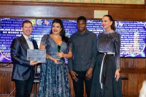 (Homem de cabelos castanhos claros vestindo um terno azul brilhante, sorrindo, com os braços alocados para trás do corpo ao lado de uma mulher branca, com vestido de gala na cor azul, sorrindo e maquiada. Ela segura um troféu de vencedora feito de murano